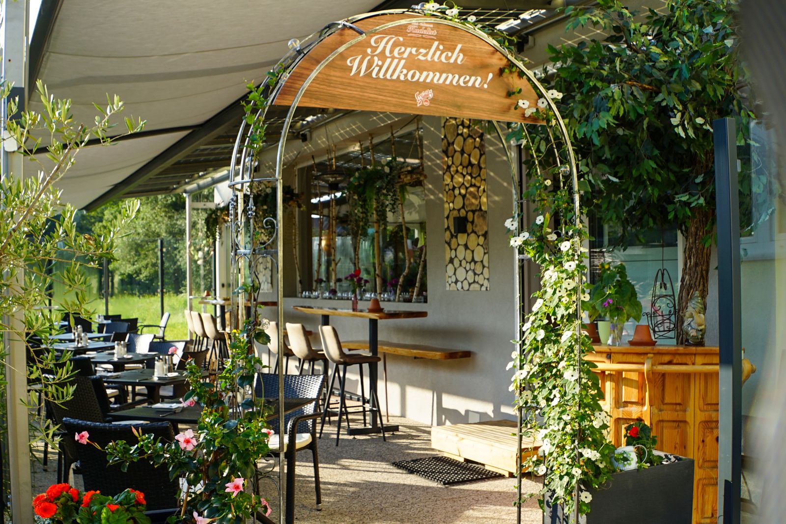 Eingang zum Gastgarten mit Rosenbogen und „Herzlich Willkommen“-Schild